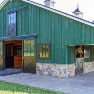 Green barn with sliding barn doors, Dutch doors, Dutch windows, and mesh stall grills against stone base.