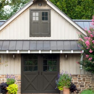 Barn with dark sliding hay loft doors, framed by plants and pink blossoms, showcasing stylish entry features.
