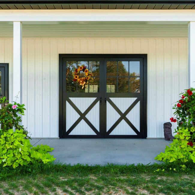 Black-trimmed entry and carriage doors on white exterior, surrounded by vibrant flowers and rocking chairs.