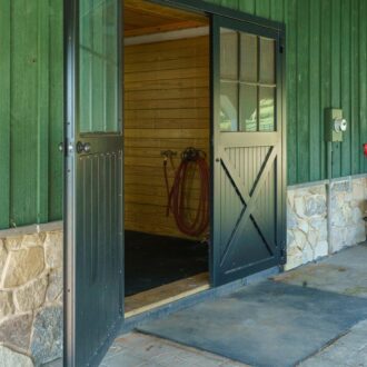 Black carriage barn door on green wall, featuring window panels, ideal for stables; customizable finishes available.