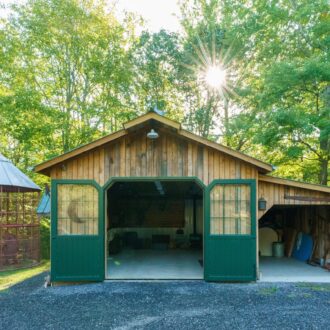 Rustic barn with green carriage doors, open to reveal spacious interior. Side structure features wood storage and large windows.