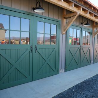Green carriage doors with glass panes, part of barn with wooden trim. Includes iron hardware and multiple finish options.