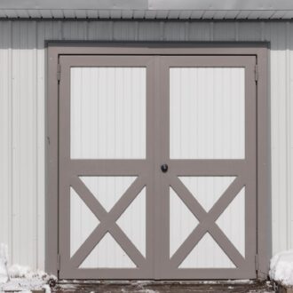 Grey and white carriage doors on a barn, featuring crossbuck design and snow-covered ground for rustic charm.