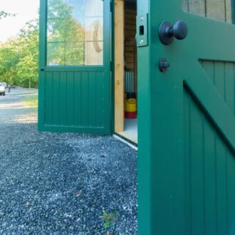 Green dutch door with glass panel, open on a wooden barn, showcasing entry door features and gravel pathway.