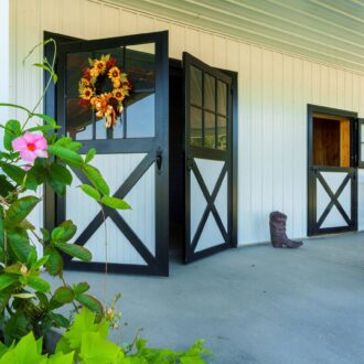 Dutch doors with black and white finish, stall grill, and decorative wreath on covered porch with plants and rocking chairs.