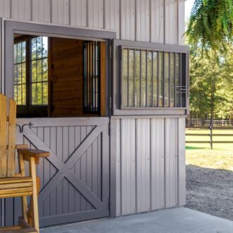 Dutch door with protective stall grill, gray finish, outdoor wooden chair, barn setting with trees in background.
