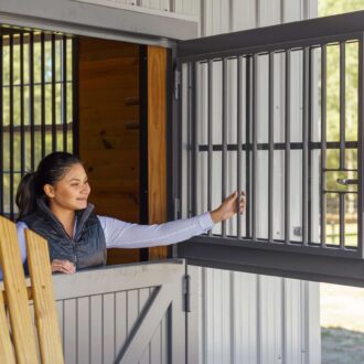 Woman opening black Dutch window with protective grill on rustic barn, showcasing versatile finish options.