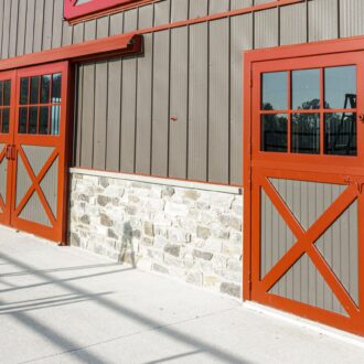 Red and gray sliding barn and Dutch entry doors on stone facade, featuring X design, glass panels, and customizable finishes.