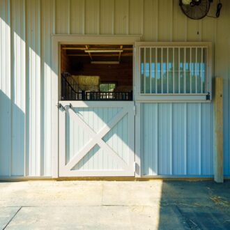 Dutch door with protective stall grills, beige finish, next to a white dutch window with bars, barn exterior.