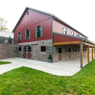 Red barn with sliding doors, Dutch windows, and Dutch horse stall doors and grills. Stone facade, metal roof, green surroundings.