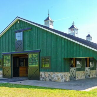 Green barn with Dutch doors, sliding barn doors, stone base, and Dutch windows under a clear blue sky.