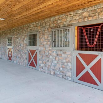 Stone barn with red and grey Dutch doors, protective stall grills, and awning windows, under wood ceiling.
