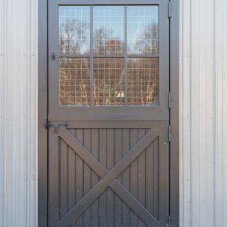 Gray Dutch door with protective stall grill on metal building, featuring a crossbuck design and customizable finishes.