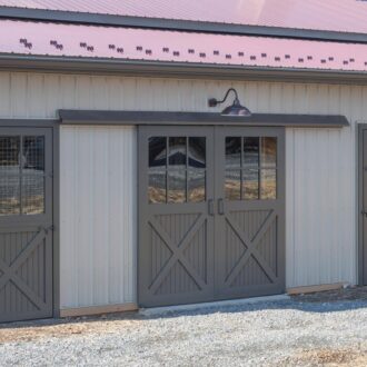 Gray sliding barn doors and entry doors with protective mesh panels on a steel building, beneath an overhanging roof.