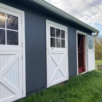 White Dutch doors with protective mesh open on gray barn exterior, featuring entry and windows in a rural setting.