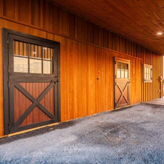 Wooden barn with Dutch doors featuring glass panes and cross-brace design, highlighted by a sunset backdrop.
