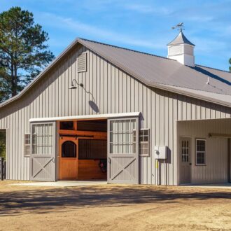 Gray barn with sliding barn doors, featuring entry doors, surrounded by trees.