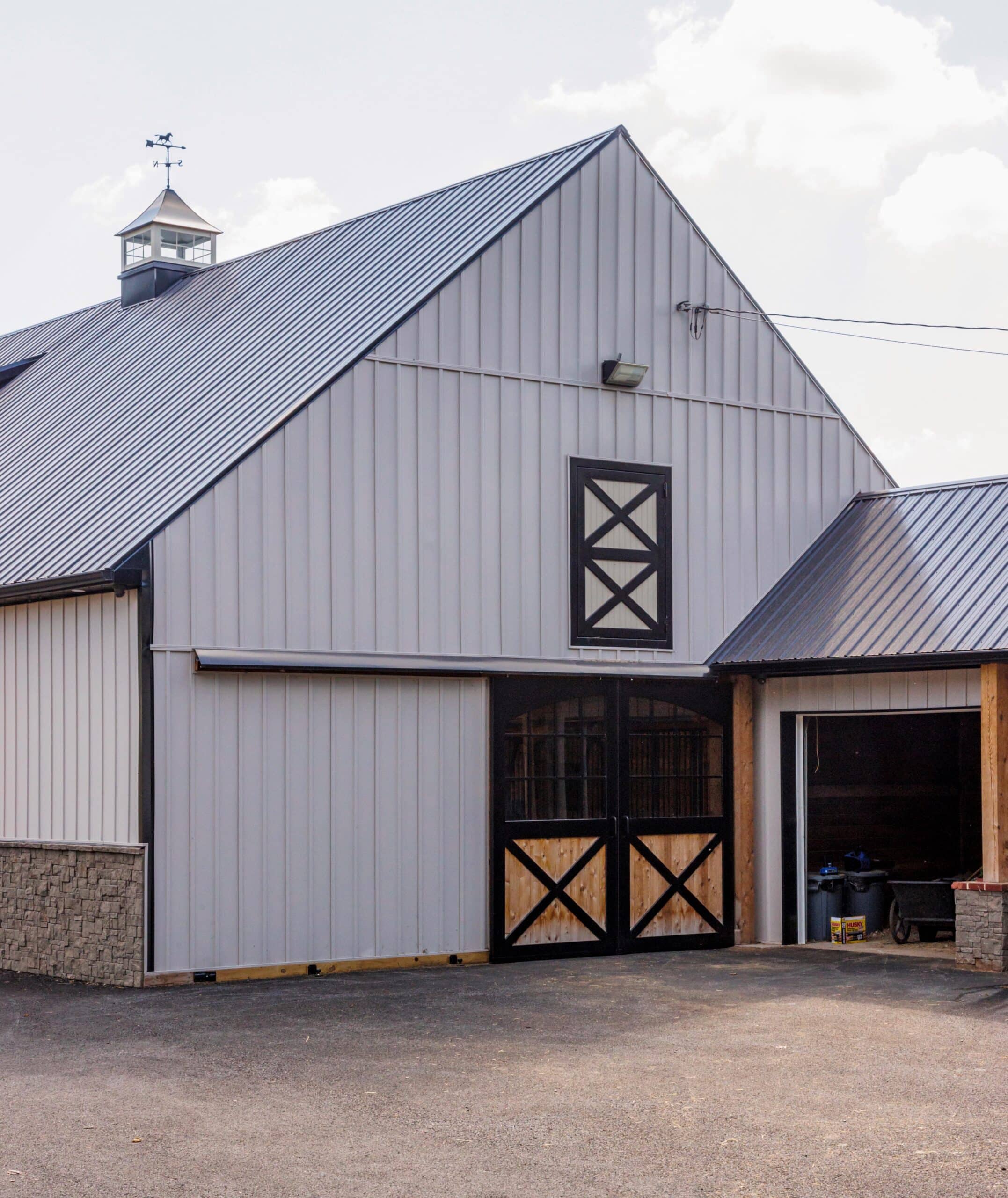 Gray barn with black sliding barn doors featuring a wooden cross pattern and a Dutch window.