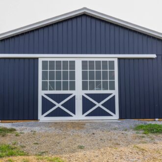 Blue barn building with white sliding barn doors featuring glass panes and crossbucks, set on gravel.