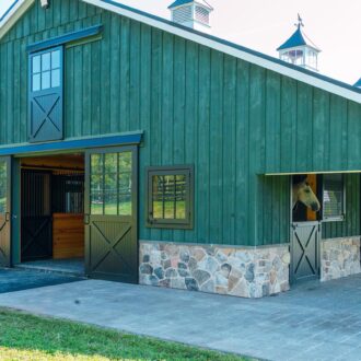 Green barn with black sliding barn doors, dutch windows, and stone accents, set in scenic countryside.