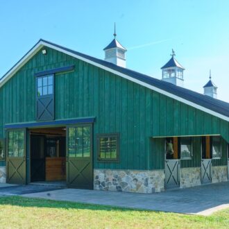 Green barn with black sliding barn doors, dutch windows, and stone accents, set in scenic countryside.