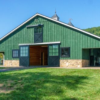 Green barn with black sliding barn doors, dutch windows, and stone accents, set in scenic countryside.