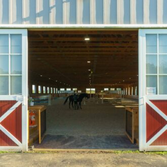Red and white sliding barn doors open to an arena with protective stall grills, showcasing a horse inside.