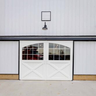 White sliding barn doors with glass panes and black trim, mounted on a barn with outdoor lights.
