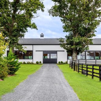 White barn with black sliding carriage doors, flanked by trees, set in spacious green landscape. Ideal for modern farm design.