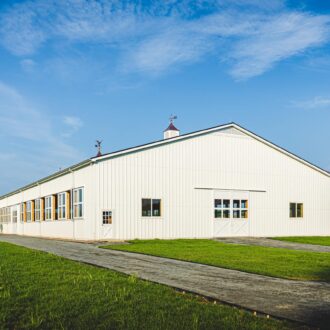 Large white arena with sliding barn doors and arena windows, surrounded by lush greenery under a clear blue sky.