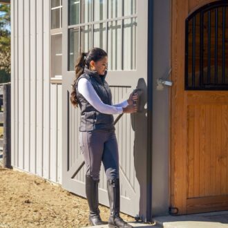 Woman opening a gray sliding barn door, next to a wood entry door with protective stall grill; outdoor setting.