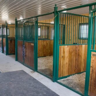 Horse stalls with green metal and wood barn doors, featuring protective grills and mesh, in a well-lit stable interior.