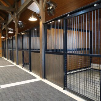 Stalls with sliding fronts, protective metal grills, dark wood finish, and overhead lighting in a barn aisle.