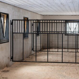 Black sliding stall door with metal grill, concrete walls, and awning windows in horse barn interior.