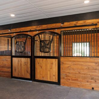 Wooden stable with sliding stall doors, black protective stall grills, and ceiling lights, showcasing natural wood finish.