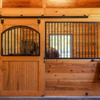 Wooden horse stall with sliding barn doors and protective grills, showcasing natural finish with durable metal features.