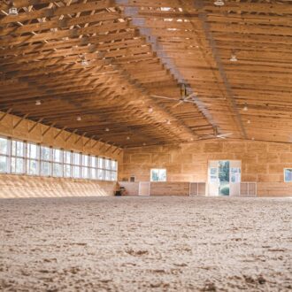 Spacious equestrian arena interior with natural light through numerous arena windows, showcasing finished wood beams.