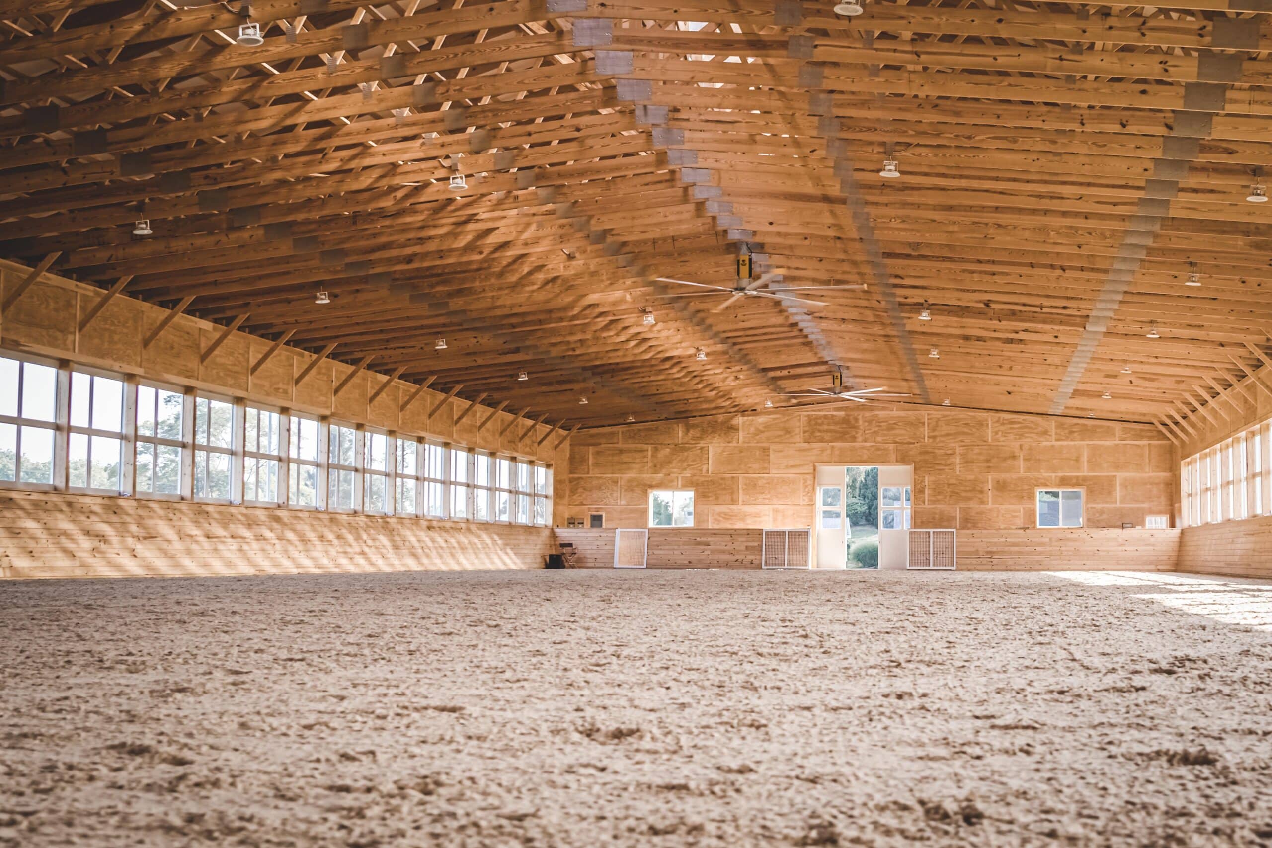 Spacious equestrian arena interior with natural light through numerous arena windows, showcasing finished wood beams.