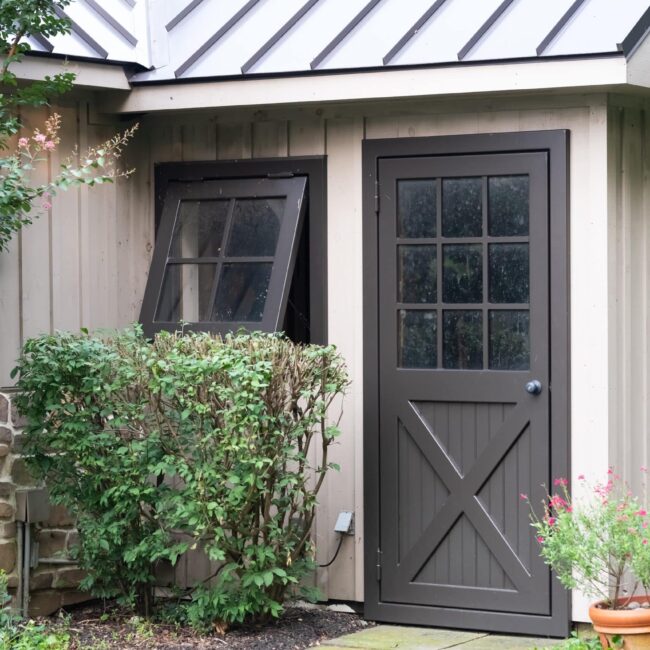 Dark gray entry door and awning windows on rustic building, surrounded by greenery, showcasing finish options.
