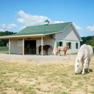 White barn with green roof, featuring sliding barn doors, dutch windows, protective stall grills, and grazing horses.