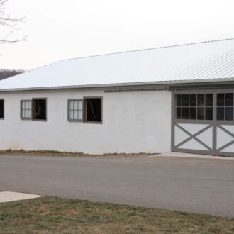 White barn with sliding doors, four Dutch windows, gray trim, asphalt drive, grass area, cloudy sky.