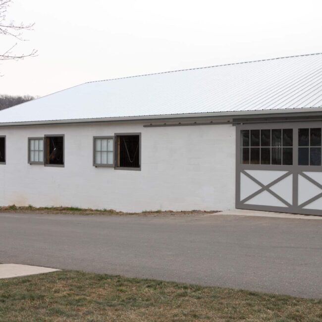 White barn with sliding doors, four Dutch windows, gray trim, asphalt drive, grass area, cloudy sky.