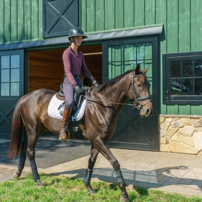 Rider on horse near green barn with sliding barn doors, dutch windows, and stone base.
