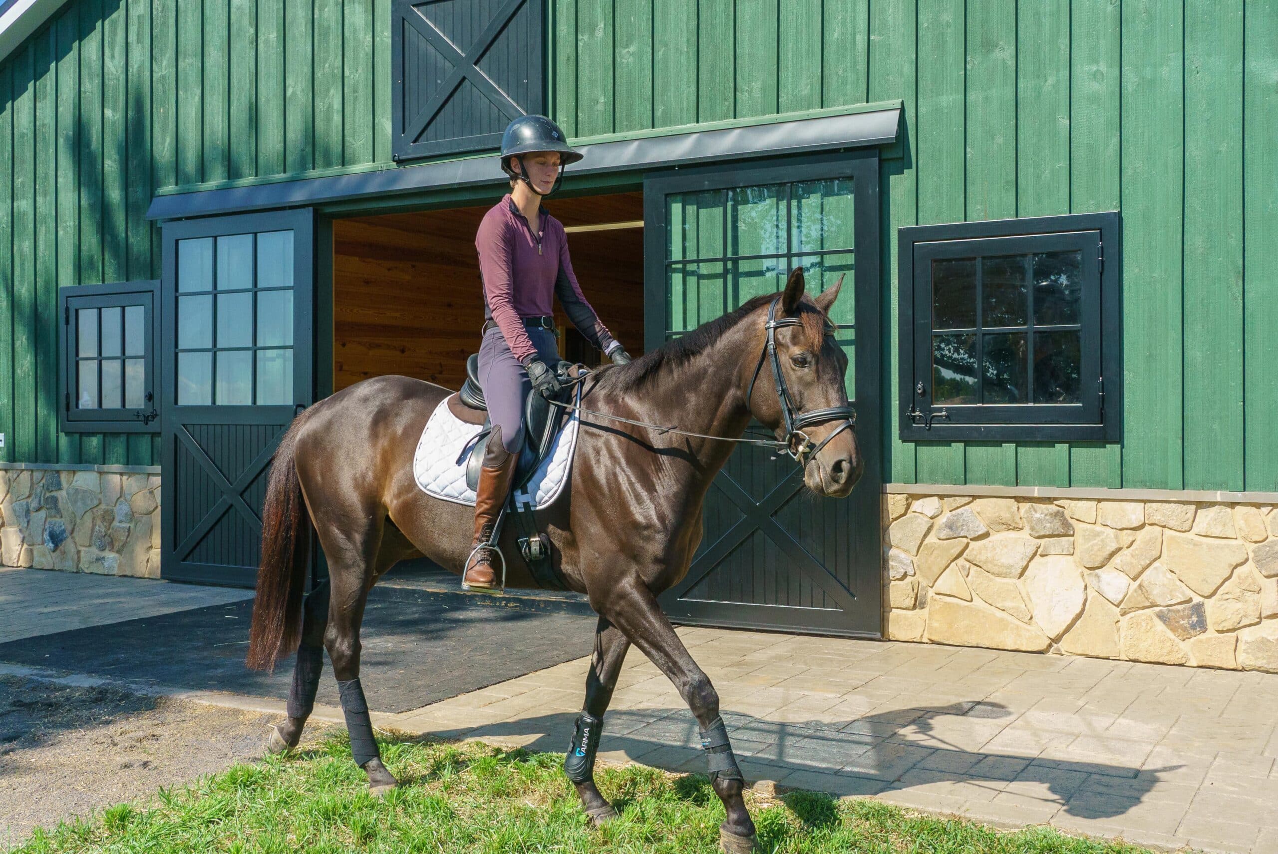 Rider on horse near green barn with sliding barn doors, dutch windows, and stone base.