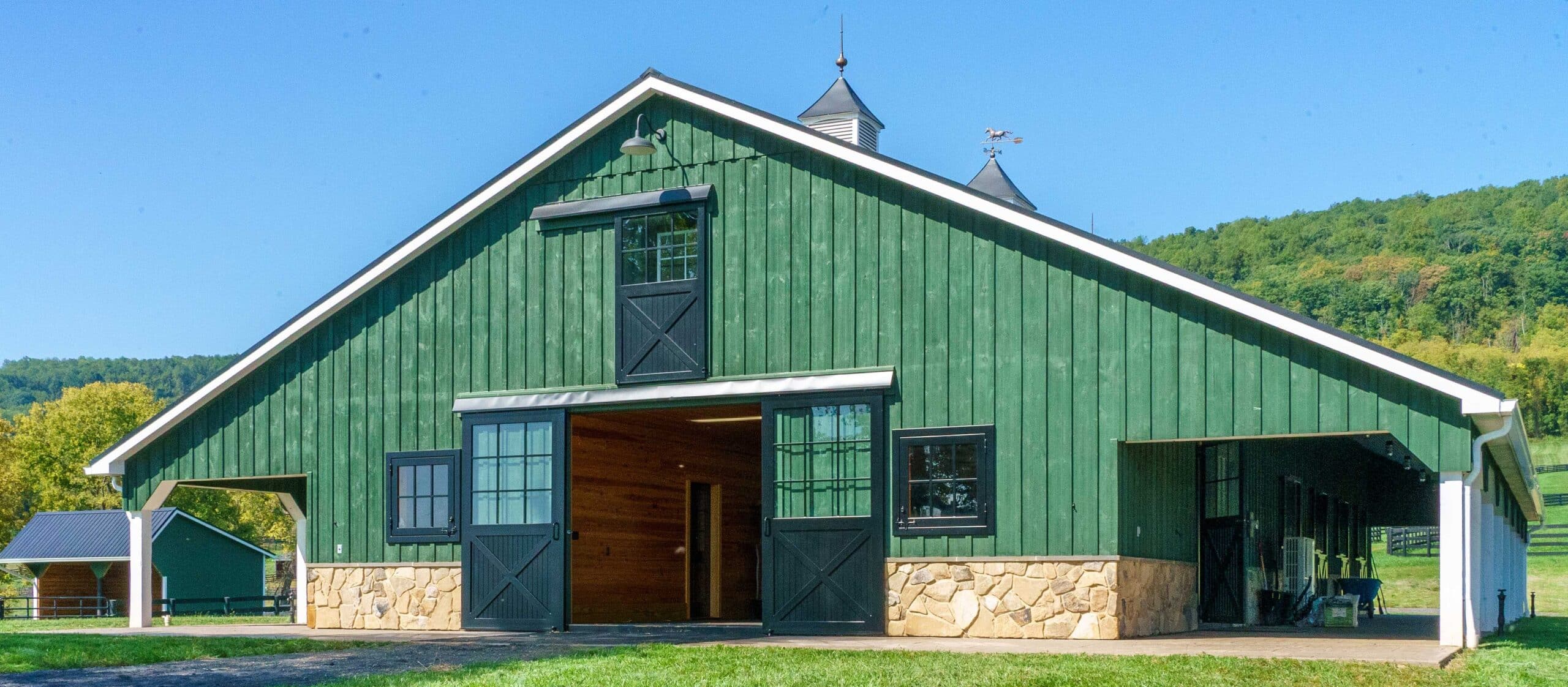 Green barn with sliding barn doors, sliding loft doors, and black Dutch windows. Stone base complements rustic design.