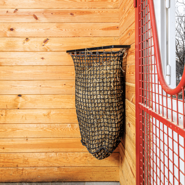 Black mesh haynet hay feeder on wooden wall. Ideal for easy feeding in stalls for horses, sheep, and other livestock.