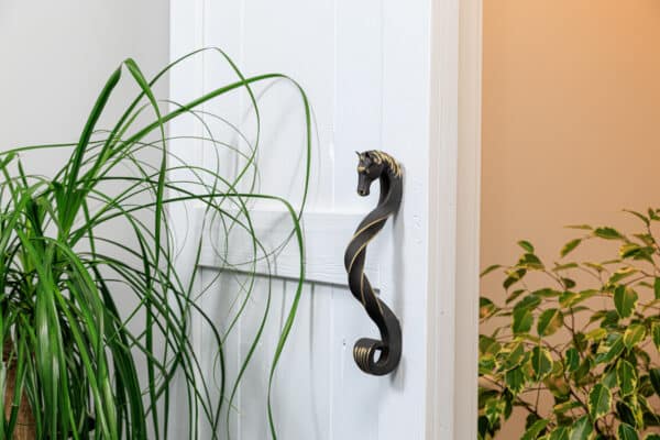 Blackened brass horse-head shaped handle on a white sliding barn door, surrounded by green plants.