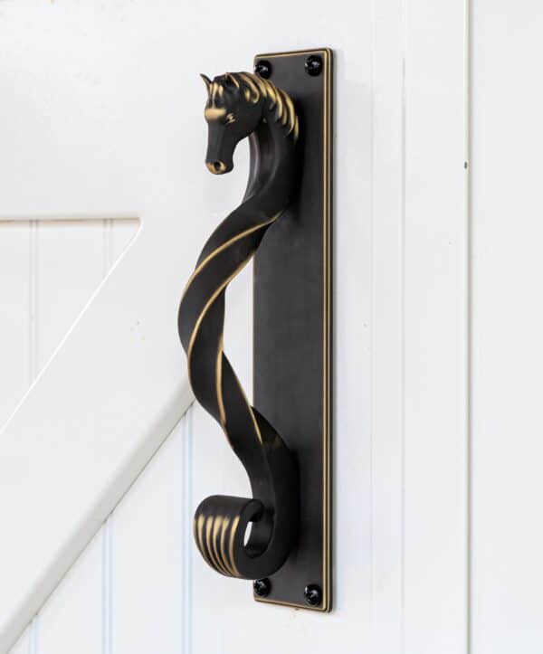 Blackened brass horse-head shaped handle with backing plate on a white sliding barn door, surrounded by green plants.