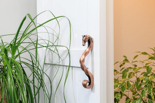 White door with decorative antique copper finish horse-handle, surrounded by lush greenery. Perfect for sliding barn doors.