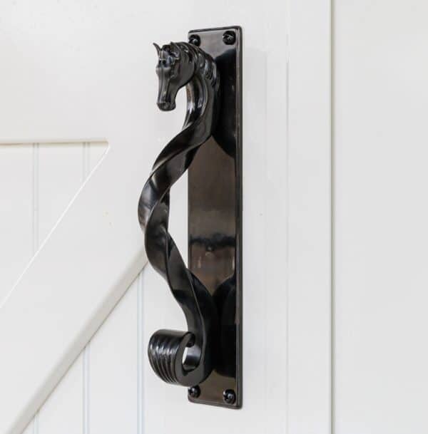 Black horse-head shaped handle with backing plate on a white sliding barn door, surrounded by green plants.